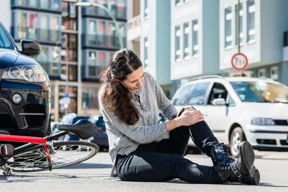 A woman sitting on the street after a bicycle accident, holding her knee in pain, with a damaged bike and cars nearby in a Brooklyn urban setting.